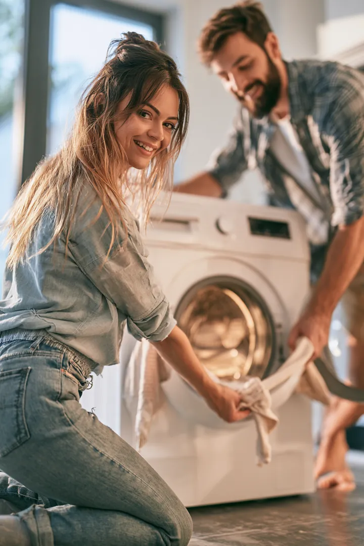 Over-the-shoulder view of a couple in their 30s sliding a new front-load washing machine into place in a modern laundry room