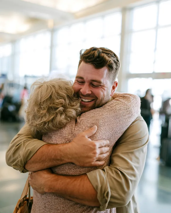 Man in his 30s hugging his mother in a bright airport arrivals hall, both beaming after a family trip covered by a $1,500 loan