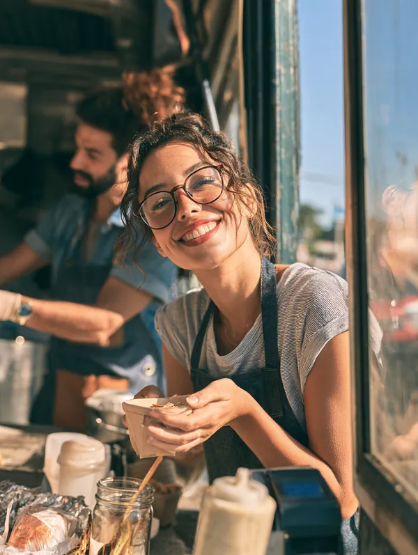 Food truck owner in her 30s and her helper opening the service window for the lunch crowd on a bright city street, a business a $5,000 loan can support