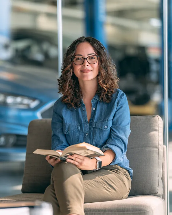 Woman in her thirties waiting calmly with a paperback in the bright customer lounge of a modern auto shop, service bay behind the glass