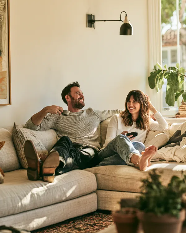 Couple in their 40s relaxing on a sectional with their feet up in a sunlit living room, calm and unburdened