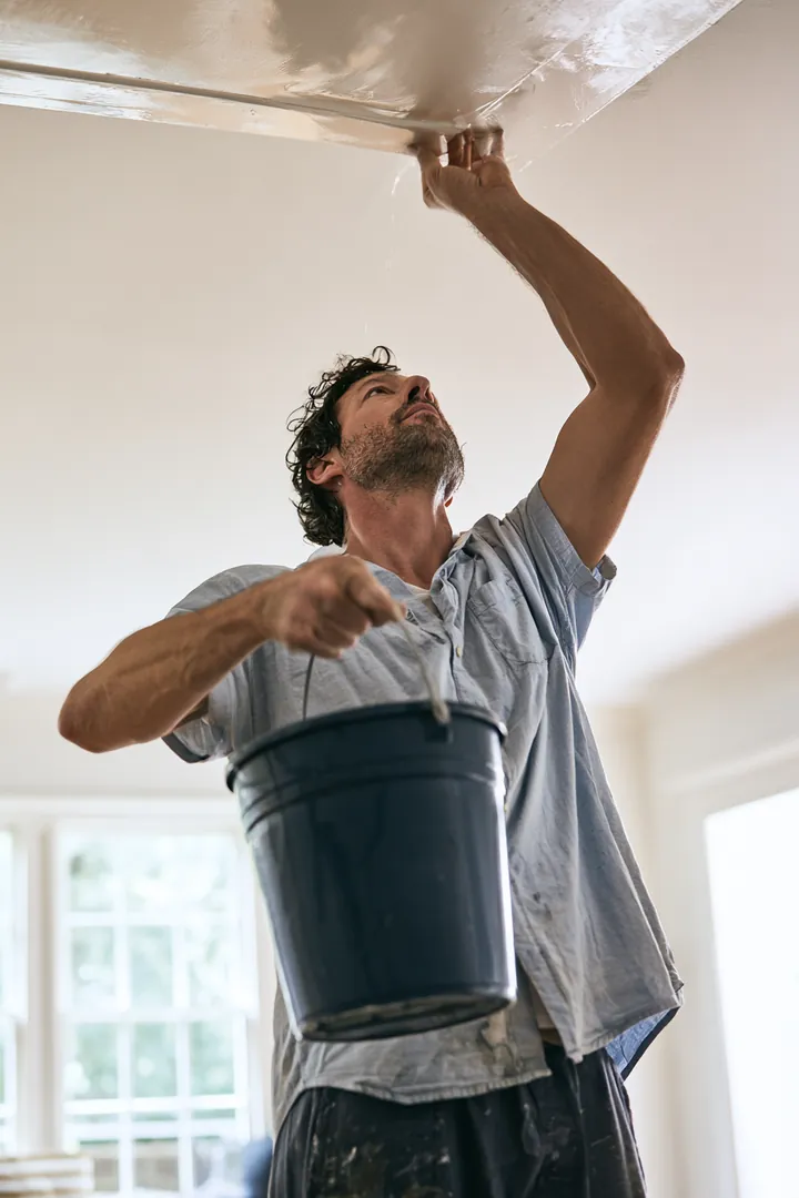 Man in his 40s placing a bucket under a ceiling drip in his living room while planning a loan instant cash request for the repair