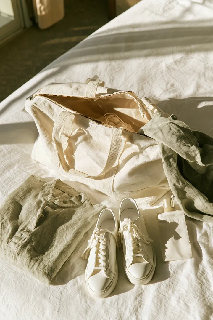 Open canvas weekender bag with white sneakers, a folded linen shirt and a travel toiletry pouch on a white hotel bedspread, a trip budgeted with a loan instant cash plan
